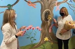 A woman wearing a white coat looks on while a woman holding a bouquet of flowers rings a bell in front of a mural of a tree.